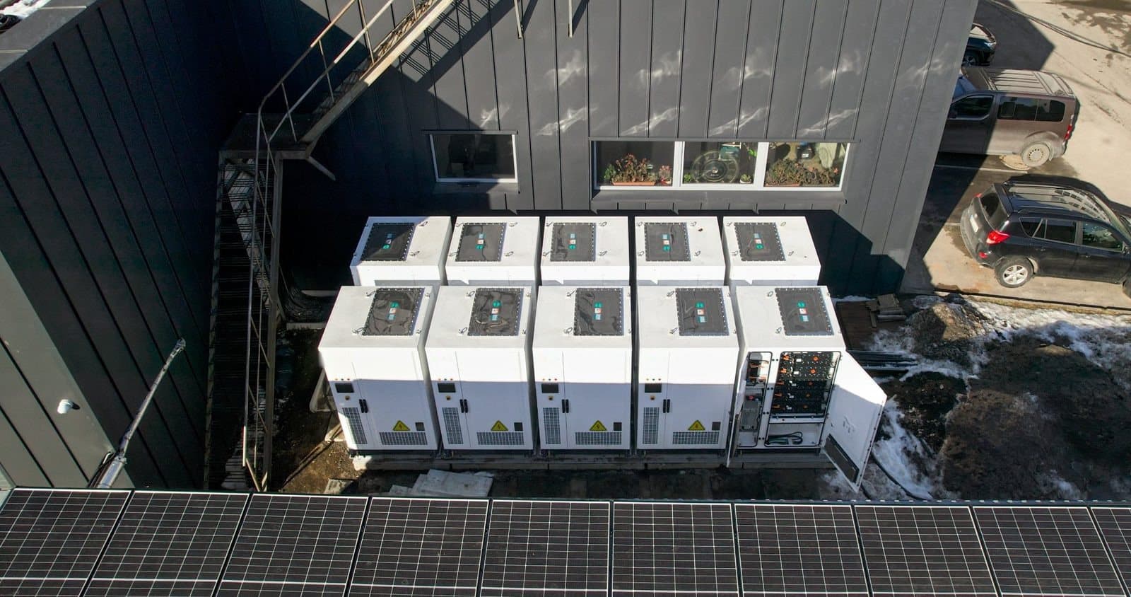 Ten battery cabinets seen from above next to a building with solar panels
